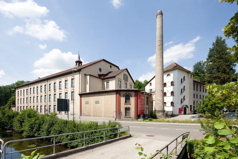 A multi-story brick and stone building with a tall chimney stack, surrounded by trees and a river or stream. The building appears to be an industrial or commercial structure, possibly a former factory or mill.