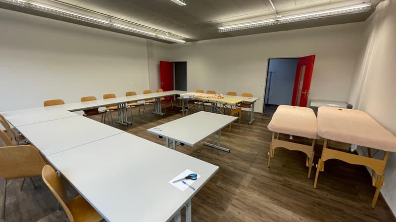 large room with white walls, wooden floor, long tables, chairs, red doors, projector