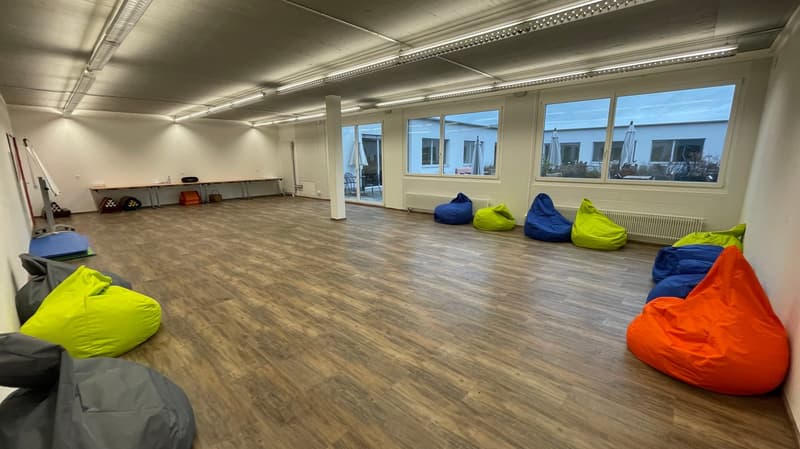 Empty room with wooden floor, white walls, large windows, bean bags, table, radiators, and ceiling lights