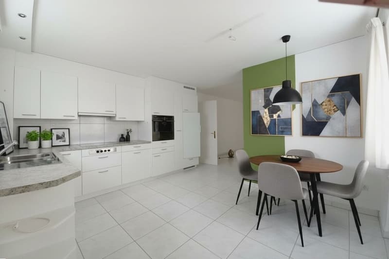Modern kitchen with white cabinetry, tile flooring, central island with sink, black appliances including an oven, a dining area with a round wooden table and chairs, green accent wall, artwork on the wall, and a window.