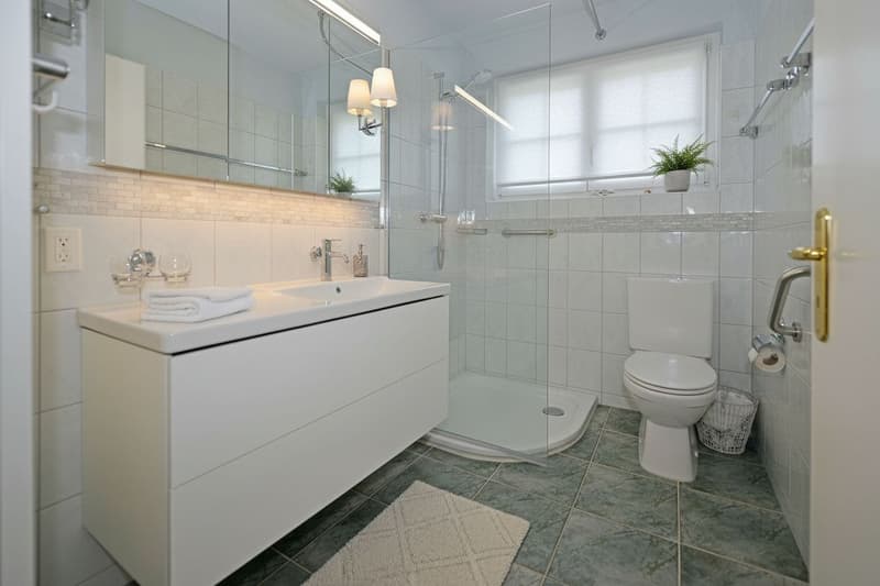 modern bathroom with white tile, white cabinet, walk-in shower, toilet, countertop with towel, power outlet, mirror with mounted lights