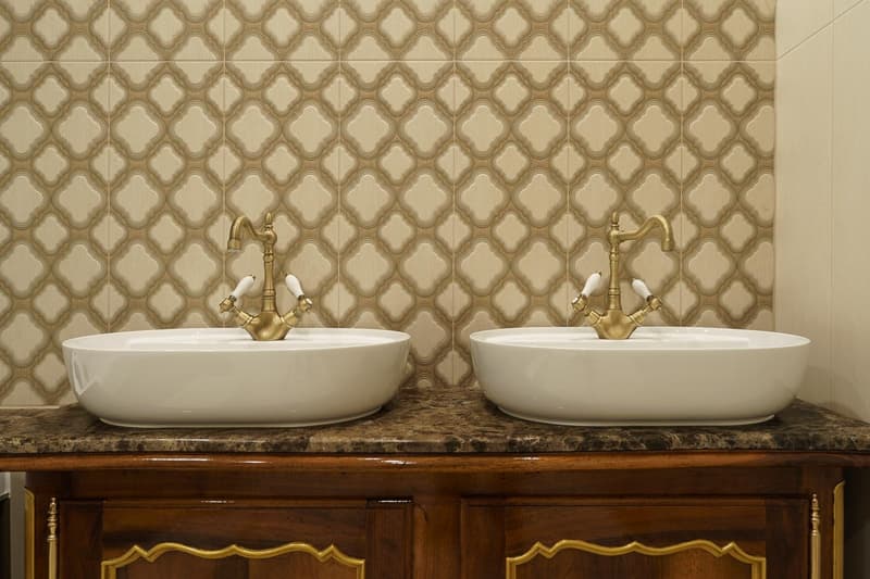 Bathroom: two basins, gold faucets, wooden cabinet, marble countertop, decorative tiles