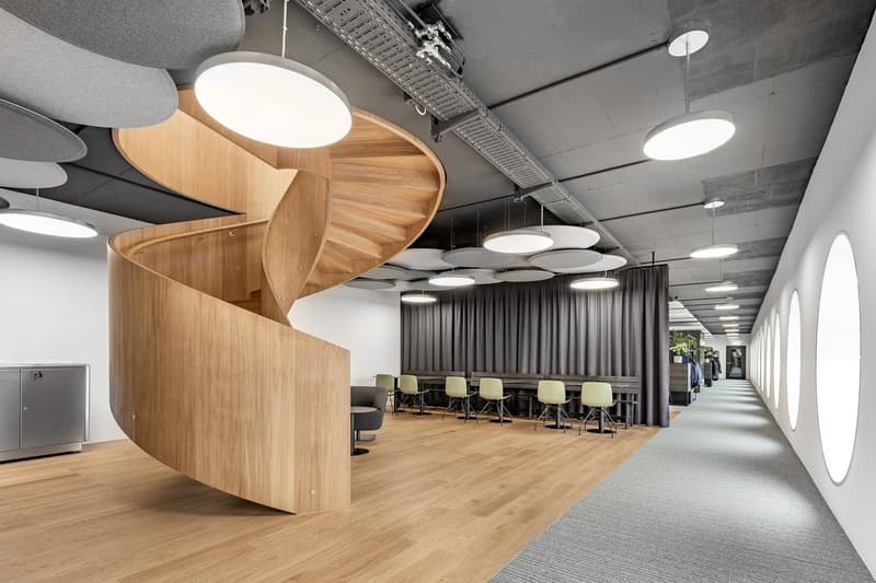 The image shows a modern and open office space with a wooden spiral staircase as a central feature. The space has a minimalist and industrial design, with exposed concrete ceilings, large windows, and circular lighting fixtures. The office area has a long