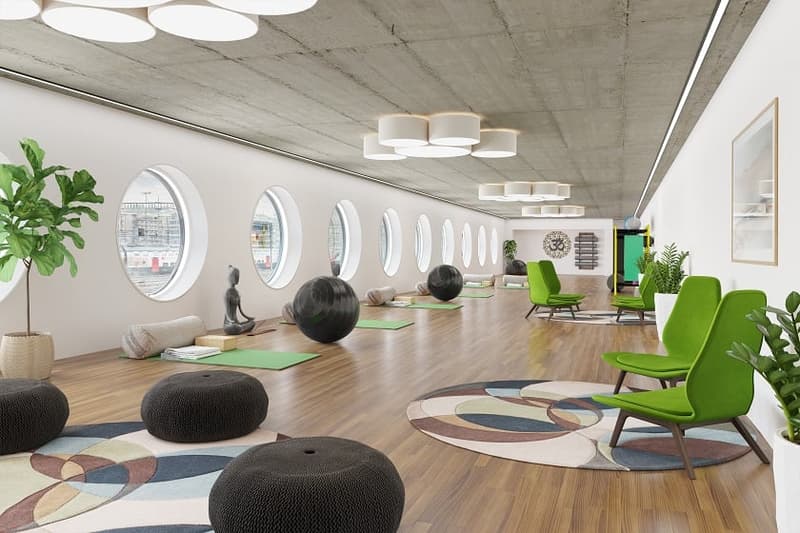 Large open space with hardwood floors, concrete ceiling, oval windows, green chairs, yoga mats on the floor, and decorative plants