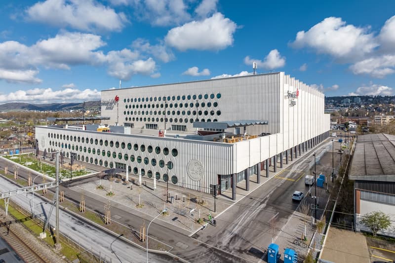 Large white industrial-style building with a distinctive circular pattern on the facade, surrounded by a paved area with some vehicles and pedestrians. The building appears to be a commercial or industrial facility located in an urban setting with mountai