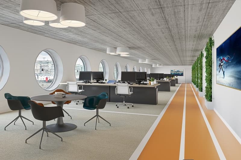 Open-plan office with rows of desks and computers, large windows, concrete ceiling, green wall feature, and a dining table with chairs