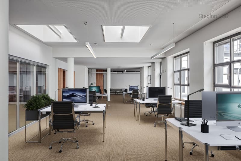 Office space with carpet flooring, modern office desks, multiple monitors, LED task lighting, large glass windows, office plants, and white walls