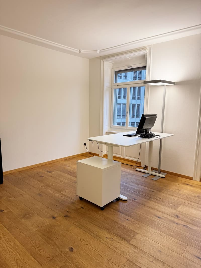 an empty room with wooden floor, white wall, a standing desk, and a lamp. There is a window with a view to the outside