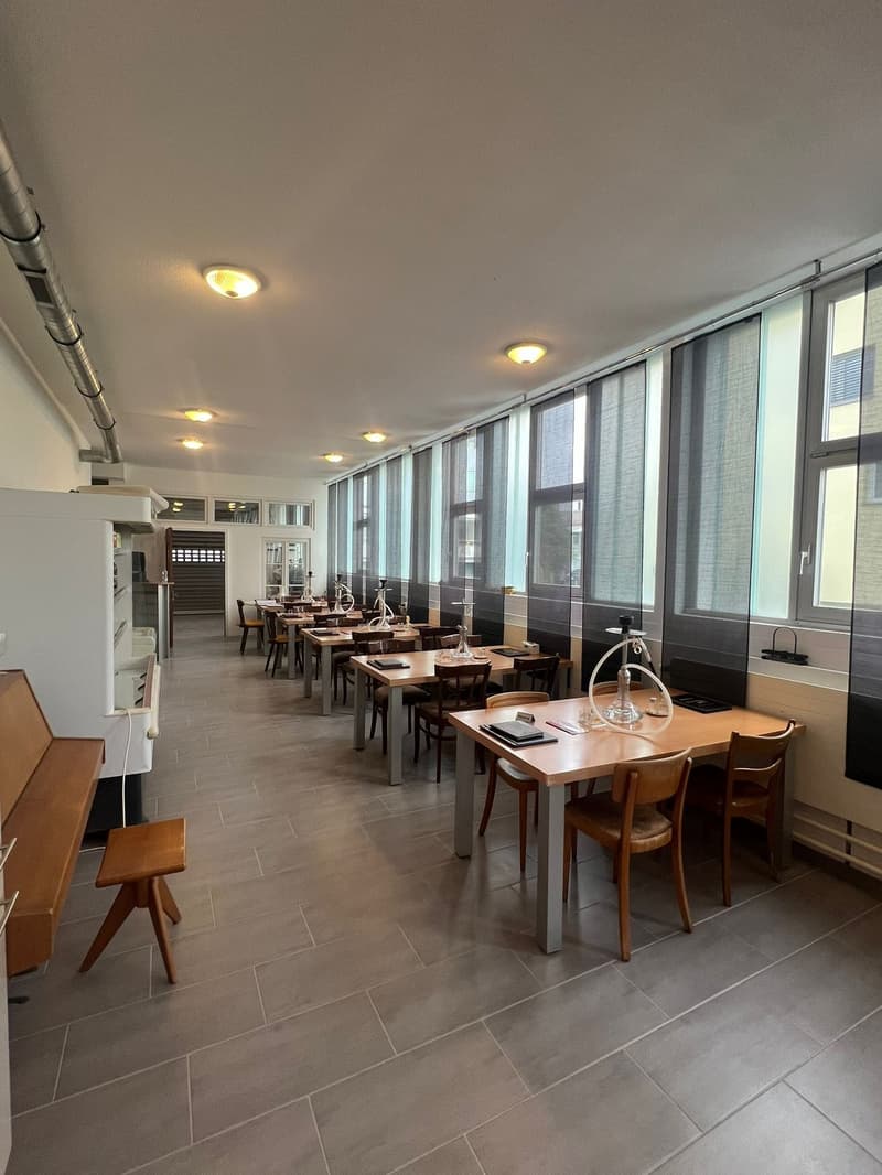 Dining hall with tables and chairs arranged, large windows with blinds, wooden bench and stool