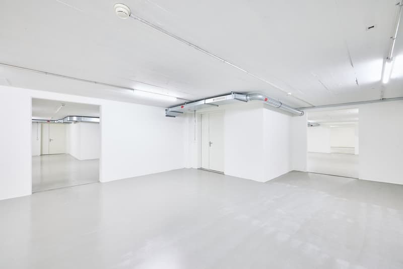 Large open commercial space with white walls, concrete floor, and overhead ventilation ducts. The space appears to be empty and ready for use as an office, studio, or other commercial purpose.