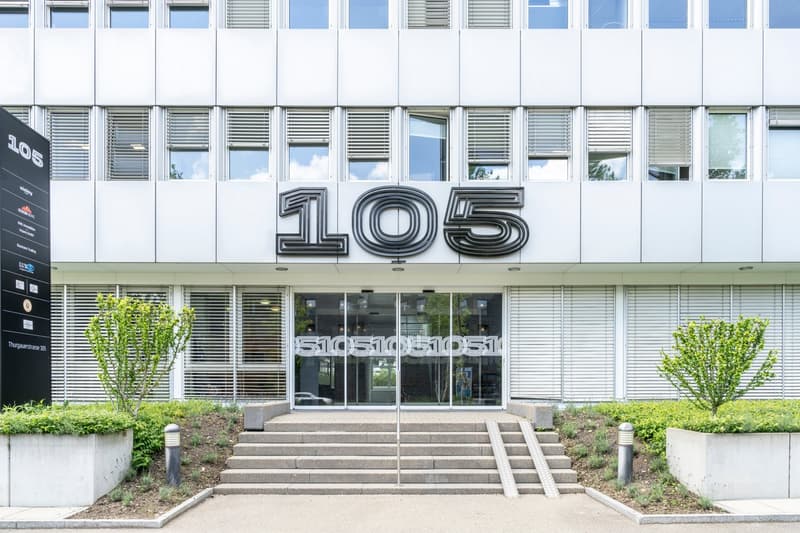 Modern office building with large windows, white facade, and prominent address number '105' displayed on the front.
