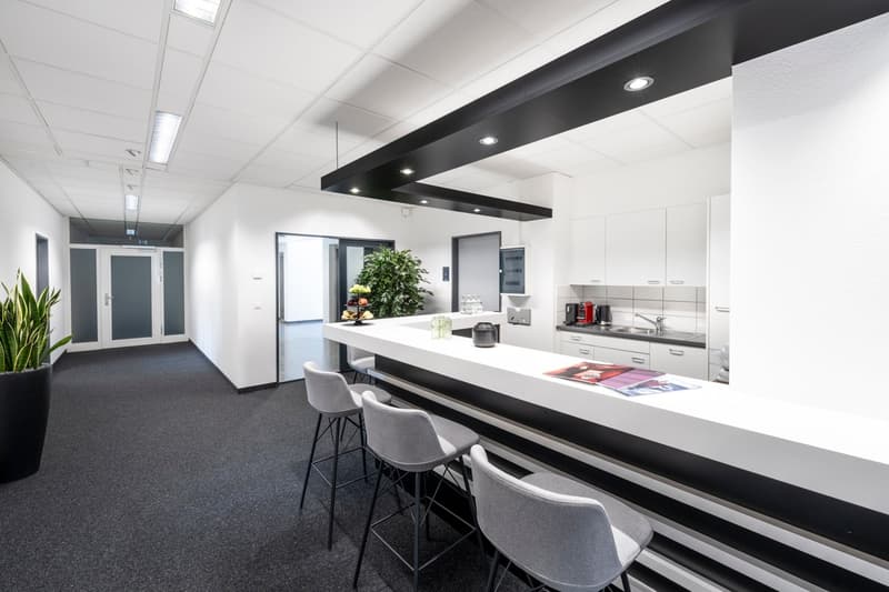 Modern office kitchen with white countertops, black accents, built-in appliances including a dishwasher, and bar seating. The space also includes a hallway with white walls and gray flooring.