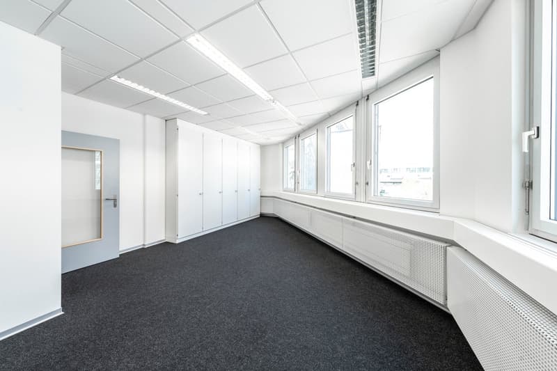 The image shows a large, open office space with white walls, a suspended ceiling with fluorescent lighting, and a carpeted floor. The room has several windows along one wall, providing natural light. There are built-in storage cabinets or lockers lining o