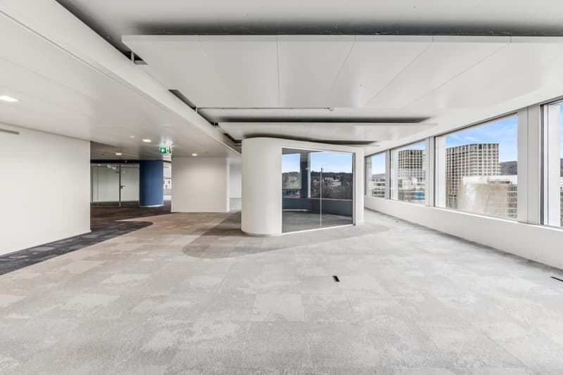 Empty office space with a view, carpeted floor, white walls, large windows, and a central cylindrical structure with a glass wall.