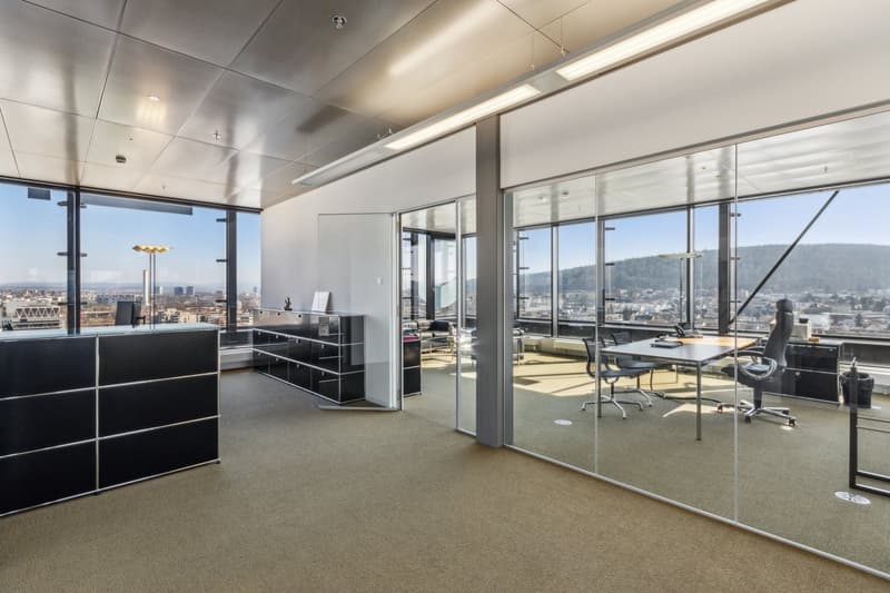 Large open office space with floor-to-ceiling windows providing a panoramic view of the city skyline. The office features a modern and minimalist design, with black storage cabinets, a conference table, and ergonomic office chairs. The lighting is provide