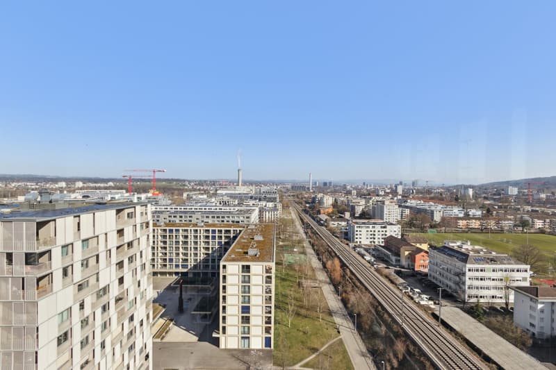 Aerial view of a cityscape with high-rise apartment buildings, railway tracks, and a mix of commercial and residential buildings. The city appears to be surrounded by hills or mountains in the distance.