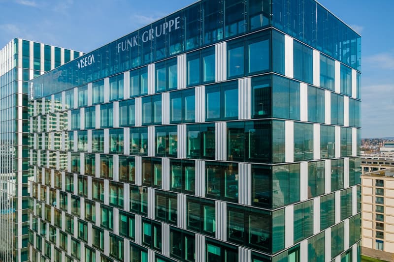 Large modern office building with glass facade, Funk Gruppe logo visible, multiple floors, parking spaces visible in front of the building