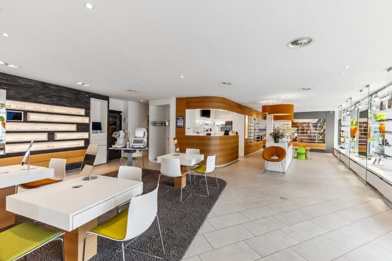 Large open-plan space with modern furniture and decor, including white tables and chairs, a curved wooden reception desk, and shelving units displaying various products. The space has a sleek and contemporary design with tile flooring and recessed lightin