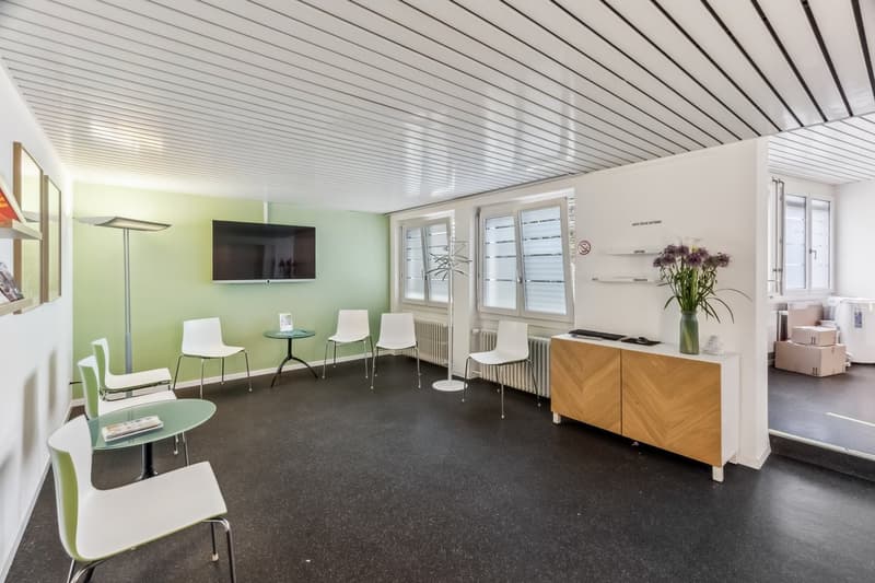Modern office space with green walls, white chairs, glass tables, TV, and a wooden cabinet with a dishwasher visible.