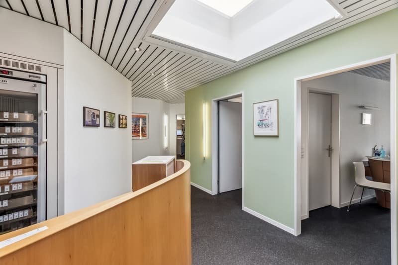 The image shows an office space with a reception area. The space has a high ceiling with a skylight, wooden reception desk, shelves with various items, and framed artwork on the walls. The walls are painted in a light green color, and the floor is covered