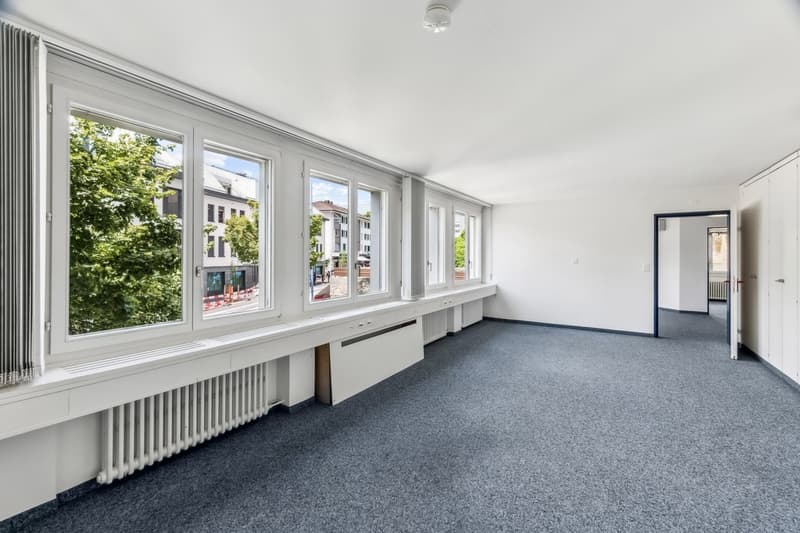 Large open office space with white walls, gray carpeted floor, and large windows overlooking a street with trees outside. The room has a radiator and a doorway leading to another room.