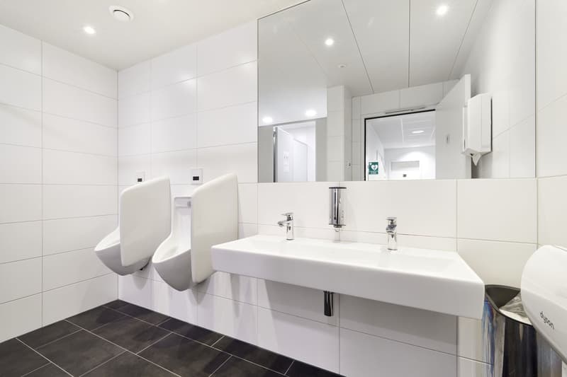 Large, modern bathroom with white tiled walls, black tile flooring, two white sinks with chrome faucets, and a dishwasher visible.