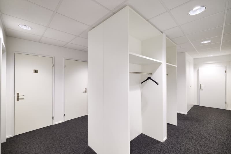 The image shows a modern office space with white walls, a carpeted floor, and recessed lighting. There is a closet or storage unit with a clothes hanger visible, and two doors leading to other rooms or areas. The overall design appears to be minimalist an