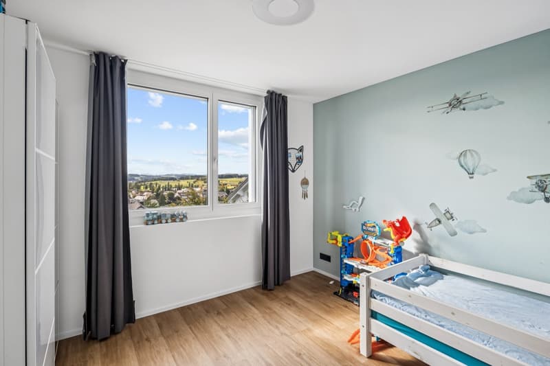 A small bedroom with a white bed, toys on the floor, white walls, wooden floor, a window with a view of the outside landscape, and a ceiling lamp.