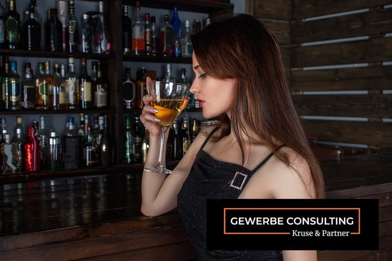 Bar counter with wooden shelf full of bottles, woman sitting on stool drinking a glass of liquor.