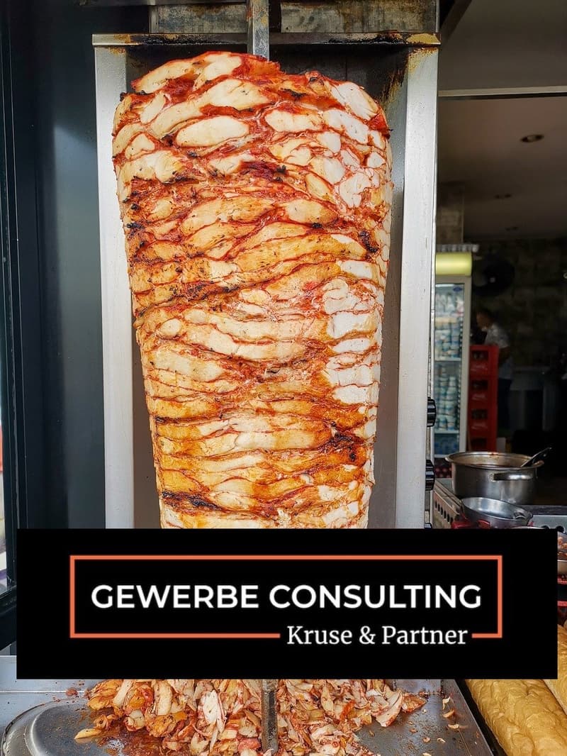 A large piece of meat, possibly chicken, is being cooked on a vertical rotisserie. The meat is sliced into thin layers and is being cooked with a red sauce. The meat is being cooked in a commercial kitchen with a stainless steel oven and a refrigerator in