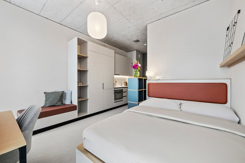 modern interior with white walls, double bed, built-in wardrobe, desk, bench with pillows, pendant lamp, mini kitchen, shelves, decorative plant