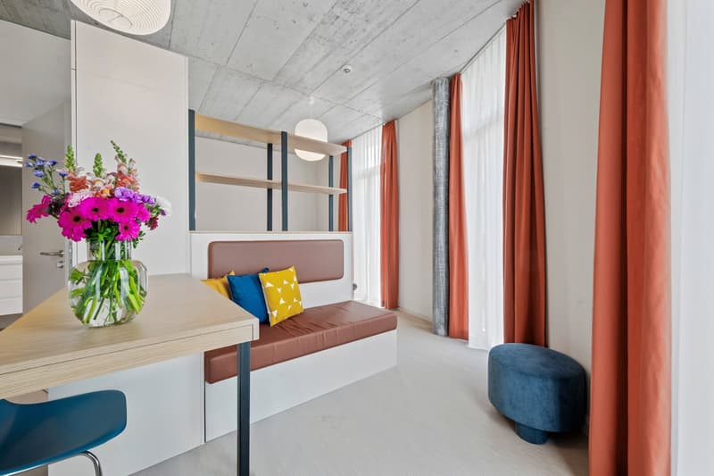 Small room with wooden bench, blue chair, small table, orange curtains, flower vase
