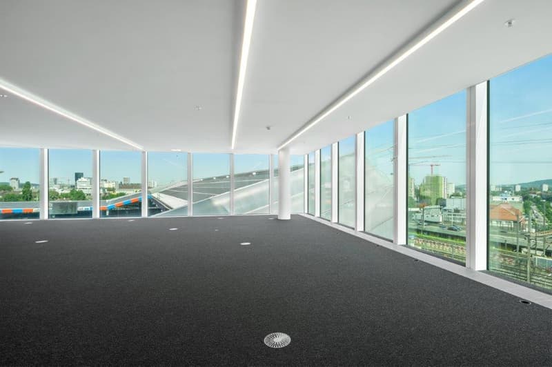 Large open office space with floor-to-ceiling windows providing a panoramic view of the city skyline. The space has a modern, minimalist design with a black carpeted floor and white walls. Recessed lighting fixtures are installed in the ceiling.