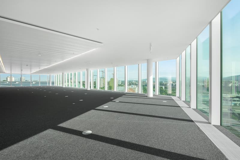 Large open office space with floor-to-ceiling windows offering a panoramic view of the city skyline. The space has a minimalist design with a grey carpeted floor and white walls. The ceiling is equipped with recessed lighting fixtures.