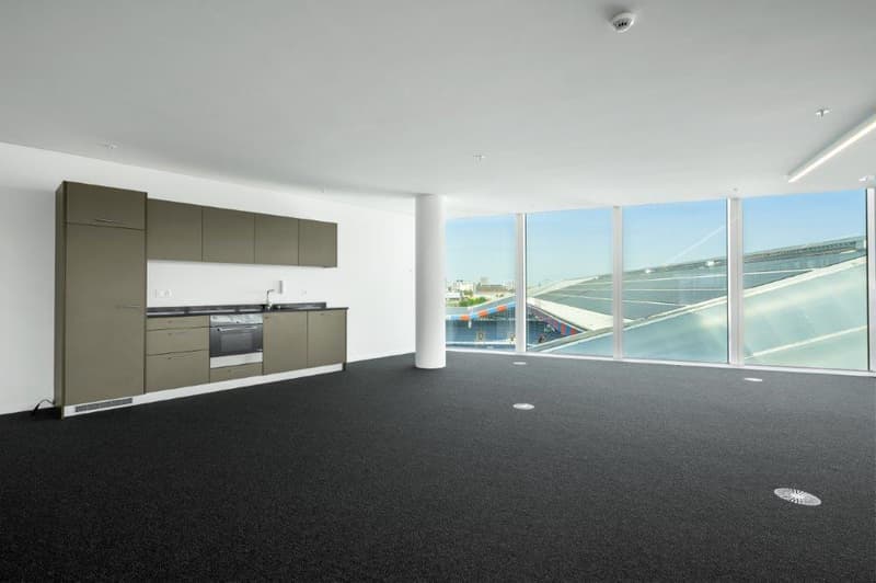 Large open-plan living space with floor-to-ceiling windows offering a panoramic view of the city skyline. The kitchen area features modern grey cabinets and appliances. The room has a minimalist, contemporary design with a dark carpeted floor.