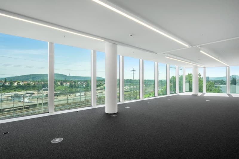 Large open office space with floor-to-ceiling windows offering a panoramic view of the surrounding landscape. The space has a modern, minimalist design with white walls, a dark gray carpet floor, and recessed lighting. The windows provide abundant natural