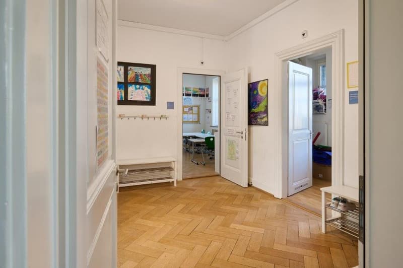 Empty room with parquet floor, white walls, and two open doors. Art displayed on the wall and a table with a chair visible in the room behind the door.
