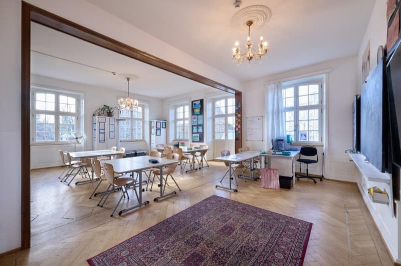 Classroom with parquet floor, large windows, tables, chairs, ceiling light, chalkboard, and plants.