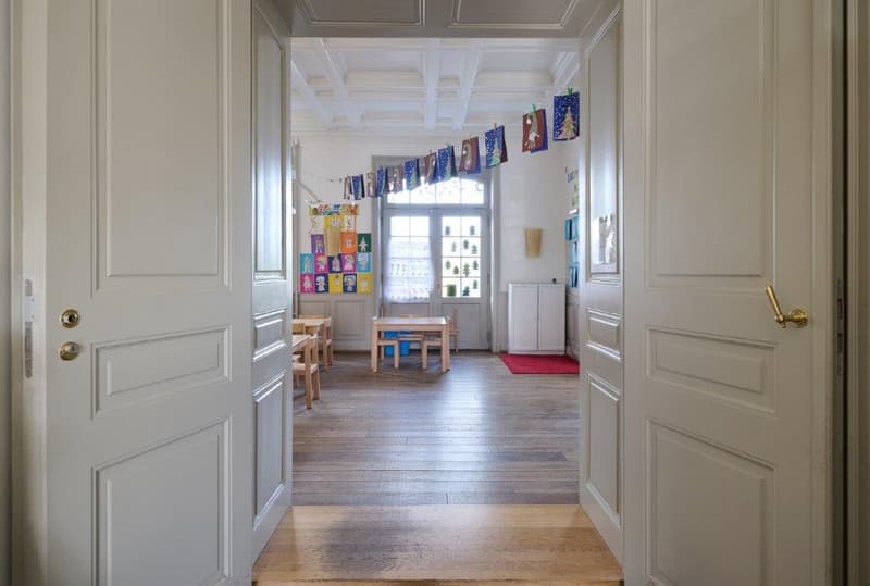 spacious room with wooden floors, white walls, decorative items on wall, windows with white frames, red carpet, wooden tables and chairs, storage cabinet.