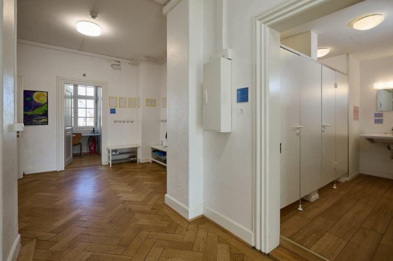 A rectangular hallway with parquet flooring, two white pillars, a large door with a mirror, and a few small tables with chairs.