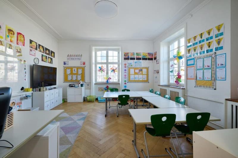 Classroom: long tables, green chairs, radiators, whiteboard, shelves with children's artwork, bulletin boards with various decorations, a television, and a radiator.