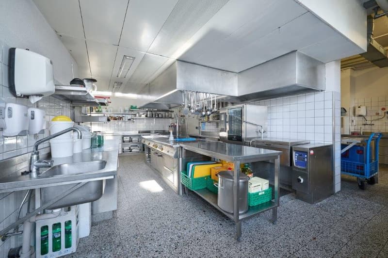 A professional kitchen with a sink, a stainless steel countertop, a stove, an oven, storage cabinets, shelves, and a dispenser.