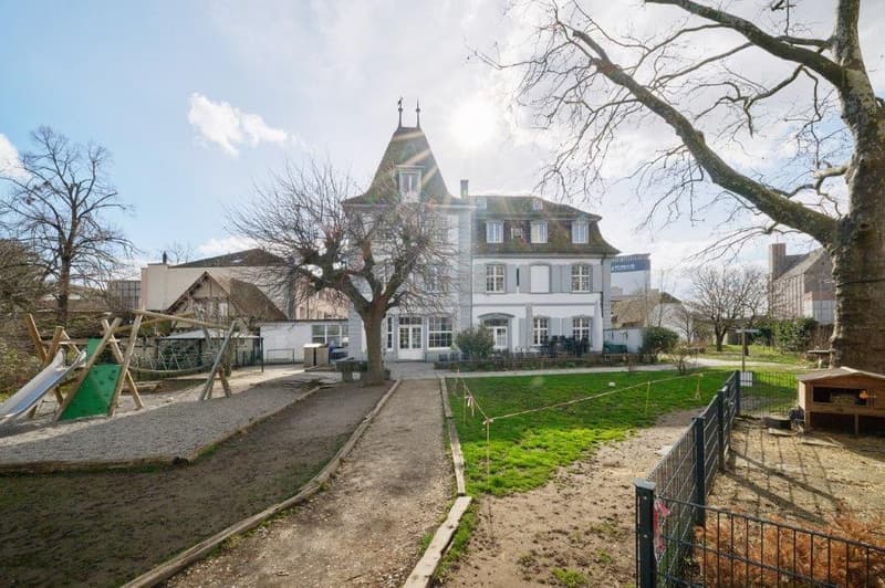 Large white building with multiple windows, brick roof, large garden, play equipment, fenced area, pathway