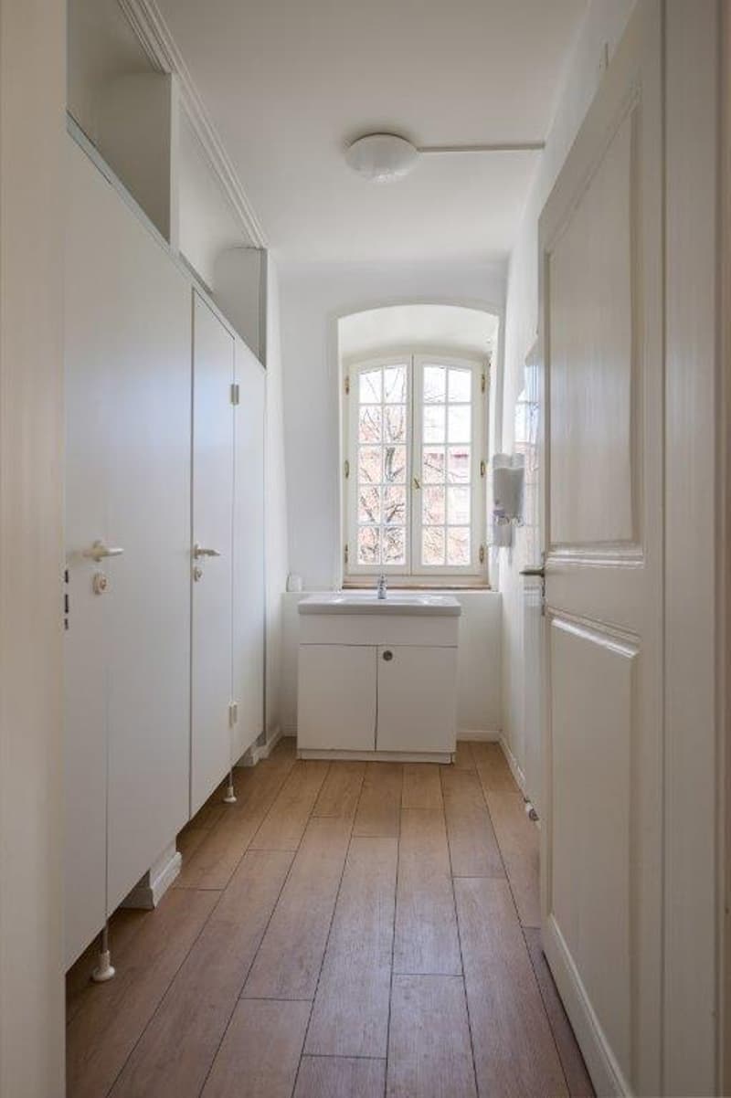 Hallway with wooden floors, white walls, glass window, white cabinet, sink, faucet, and toilet stalls.