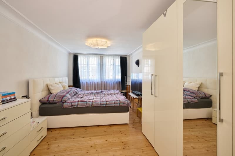 bedroom with wooden floor, double bed, white bedding, white furniture, lamps, windows with white curtains, storage box, books, desk