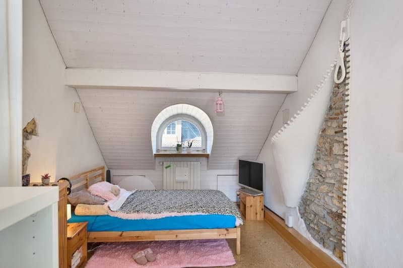 A bedroom with a wooden bed, pillows, blanket, rug, wooden nightstand, shelf, television, heater, stone wall and window.