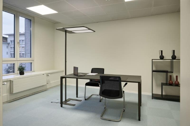 The image shows a modern office space with a desk, chair, and shelving unit. The room has large windows, a neutral color scheme, and recessed lighting. The overall design appears to be minimalist and functional.