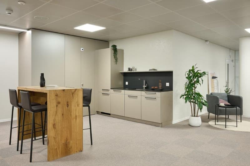 Open-plan office space with a kitchenette area, a wooden table with bar stools, and a seating area with a couch and armchair. The space has a modern, minimalist design with light-colored walls and floors.