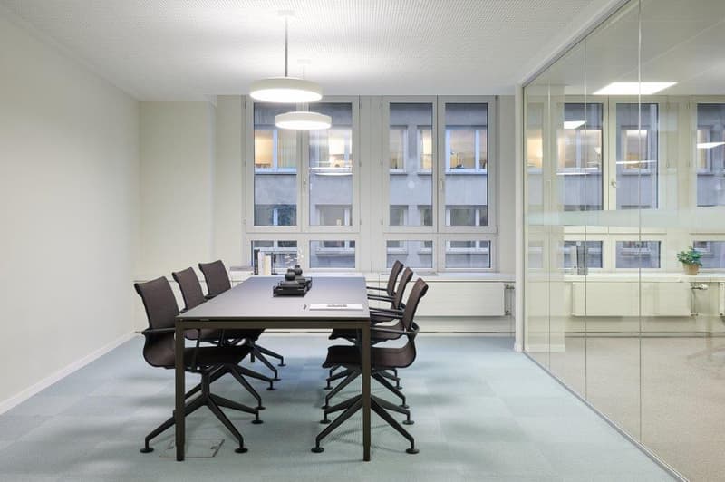 Large conference room with a long wooden table, black leather chairs, and floor-to-ceiling windows overlooking the city. The room has a modern, minimalist design with white walls and a gray carpet. There is a dishwasher visible in the room, likely for con