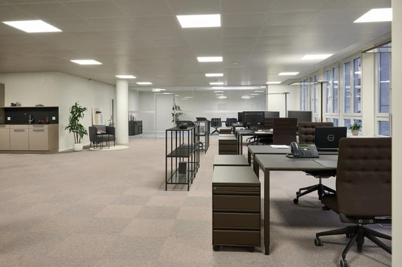 Large open office space with cubicles, desks, chairs, and computer equipment. The space has a modern and minimalist design with white walls, gray carpeting, and recessed lighting.